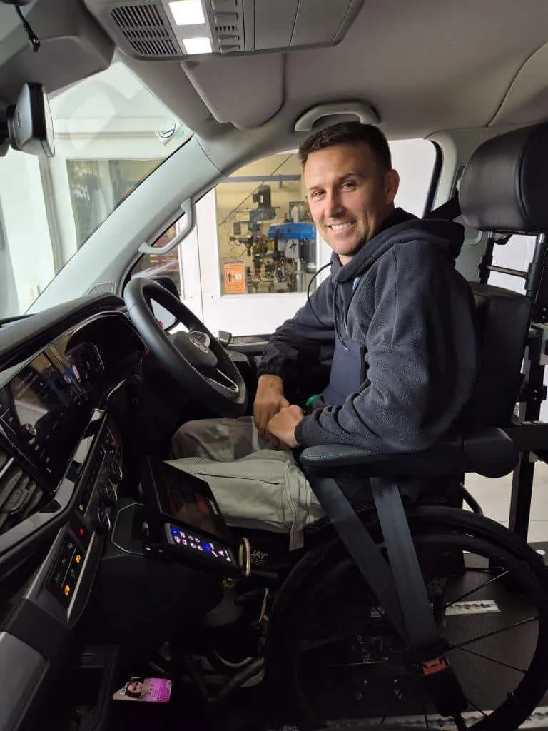 A man seated in a wheelchair-adapted driver position, smiling at the camera